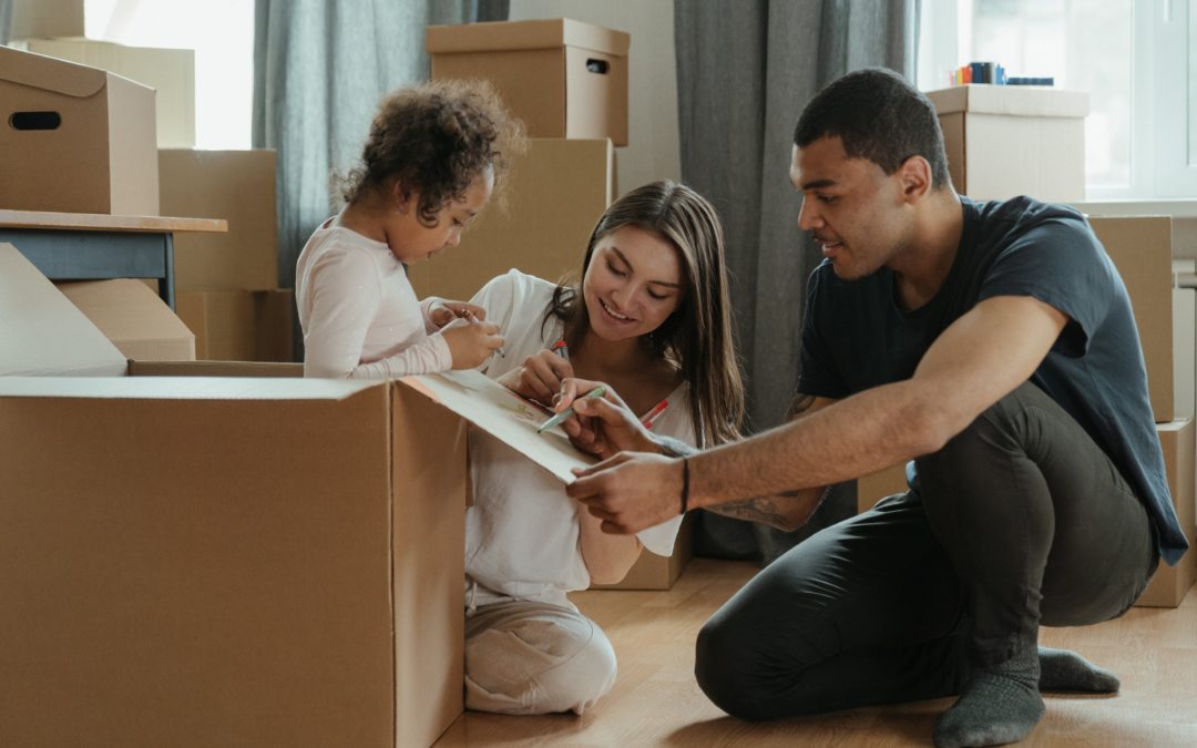 Photograph of a smiling multiracial family, a man, woman and small girl sitting on their floor at home surrounded by large cardboard boxes. The father is labelling one of the boxes using a marker pen.