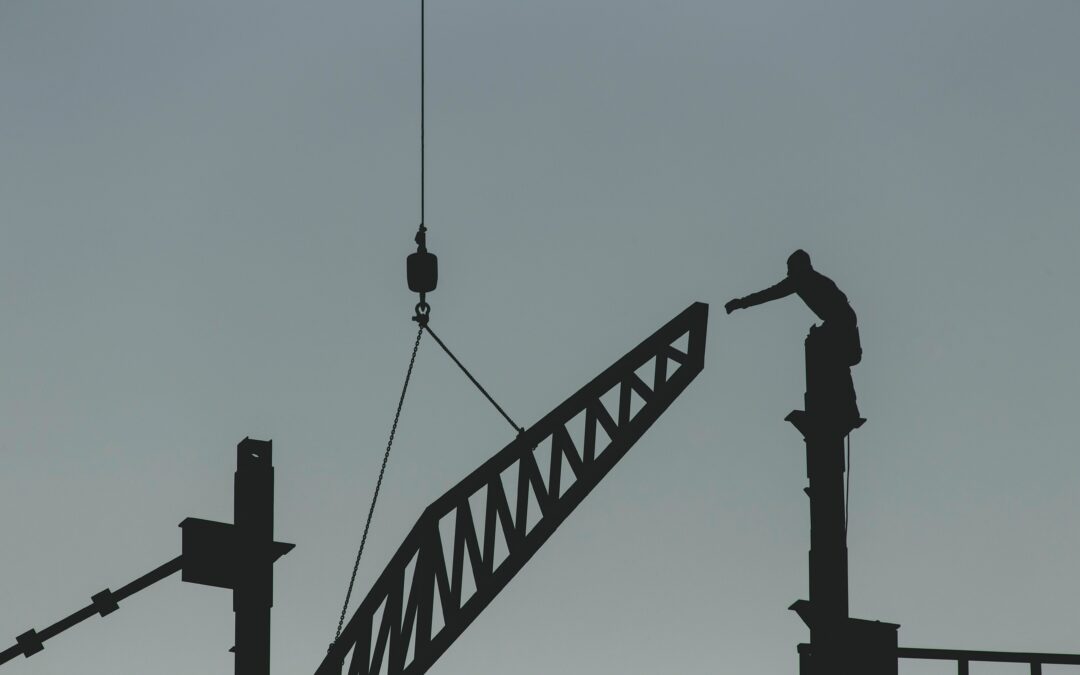 Photo of a silhouetted man standing on a high beam and stretching his hands towards a crane