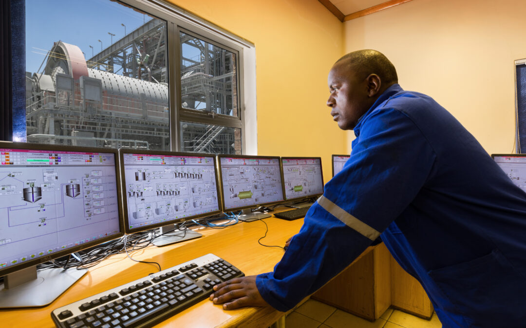 A man stands at a desk examining several computer screens. A window in the room looks out at mining machinery outside.