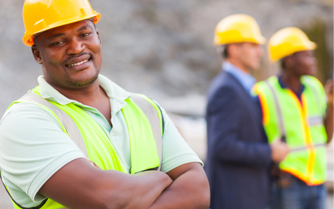 Man in a utility vest and yellow hardhat smiling at camera and standing with arms crossed, with two other blurry men in hardhats standing further away.