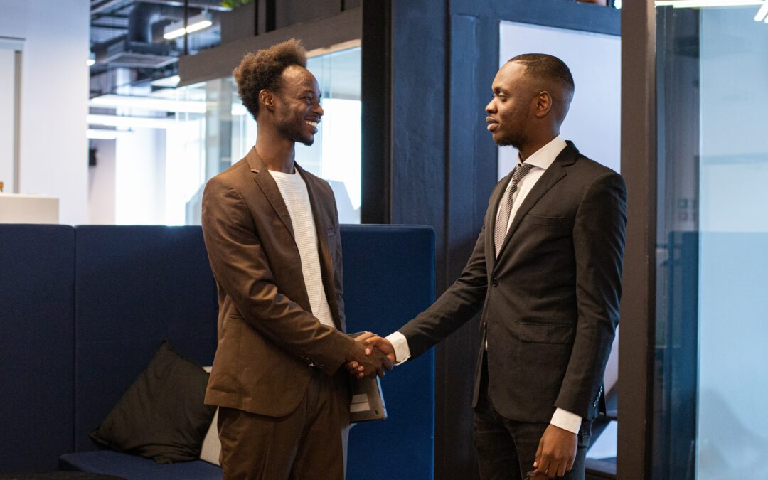 Two men wearing dark business suits standing in an office and shaking hands and smiling at one another