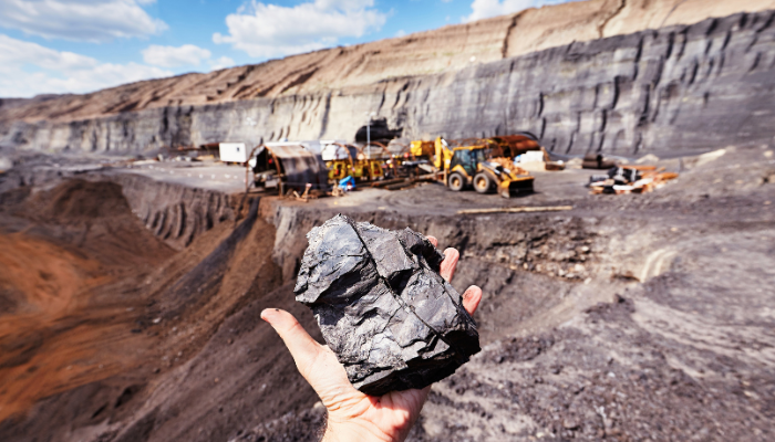 A hand holding a large piece of coal, with a mine site in the background where yellow large trucks are operating
